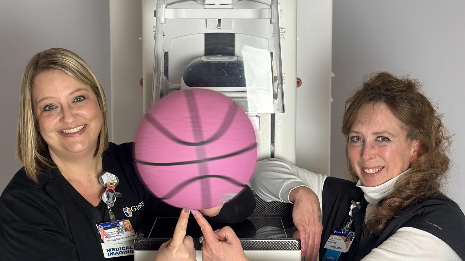 Two women in medical scrubs spin a pink basketball in front of a mammography unit.