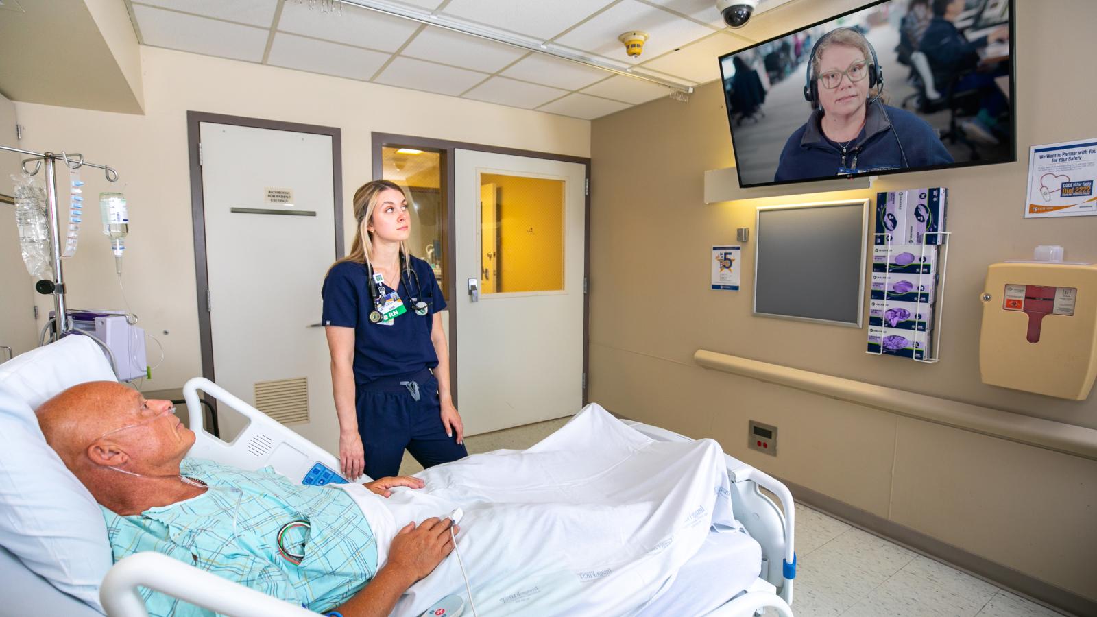 A nurse stands next to a patient in a hospital room while speaking with a virtual nurse on a wall-mounted screen.