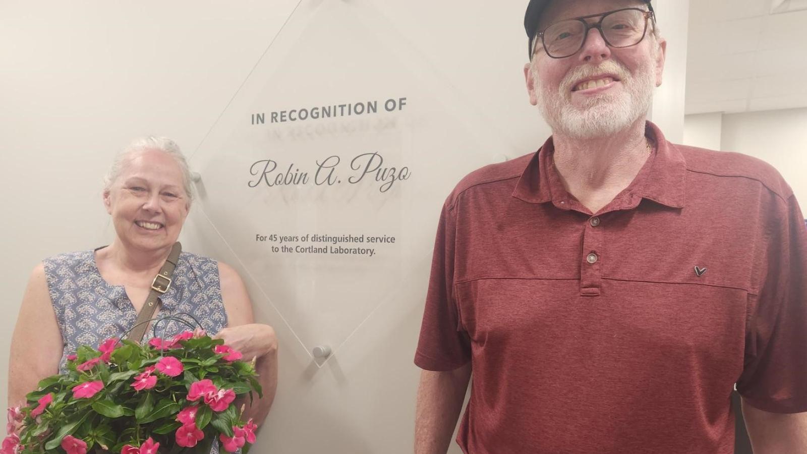 A woman holding flowers and a man pose in front of a diamond-shaped glass plaque hung on a wall.