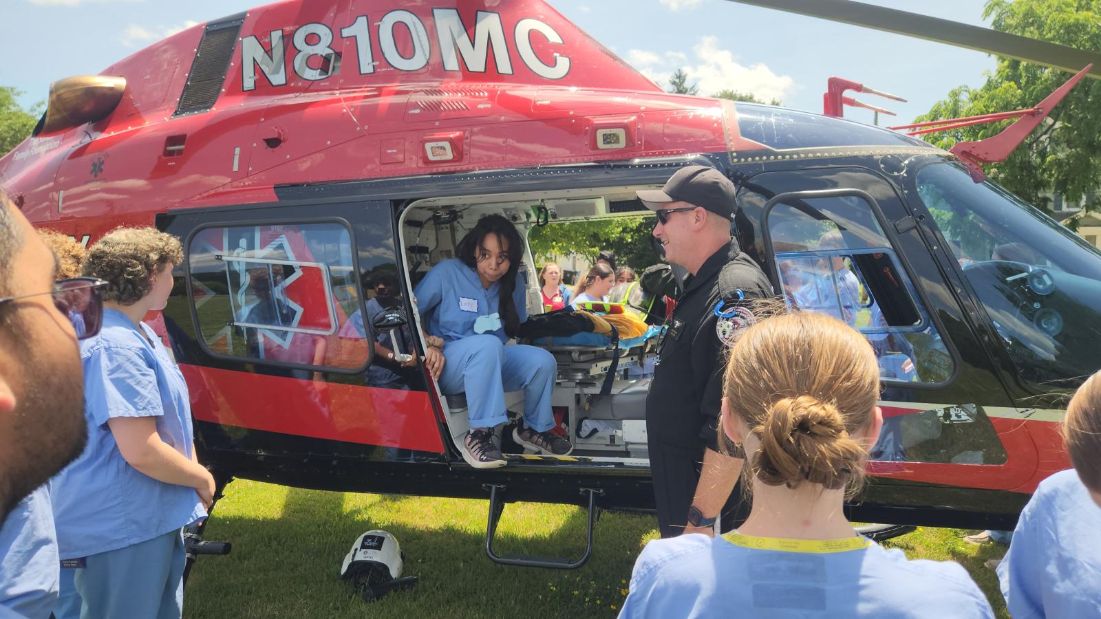 Students in medical scrubs gather around a helicopter and talk with one of the crew. One of the students is sitting in the helicopter.