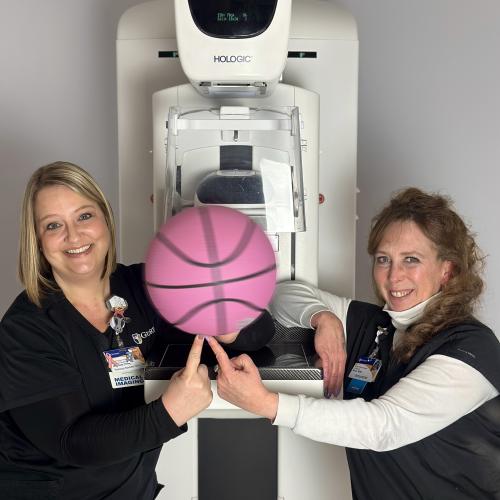 Two women in medical scrubs spin a pink basketball in front of a mammography unit.