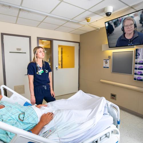 A nurse stands next to a patient in a hospital room while speaking with a virtual nurse on a wall-mounted screen.
