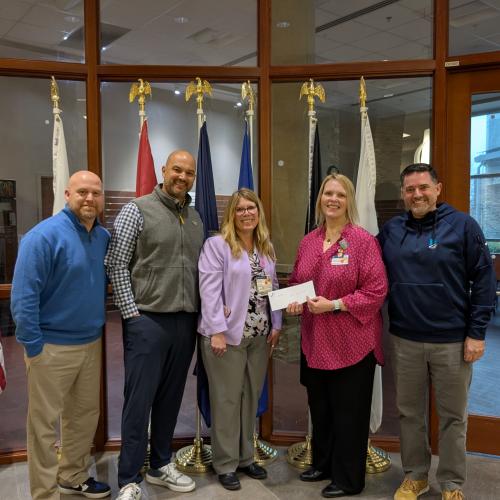A group of five individuals pose for a photo while displaying a check for a donation.