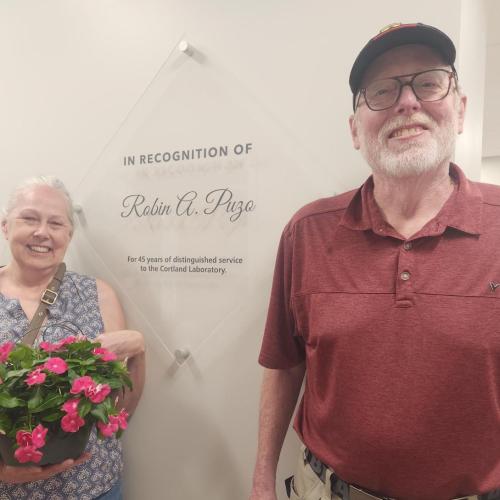A woman holding flowers and a man pose in front of a diamond-shaped glass plaque hung on a wall.