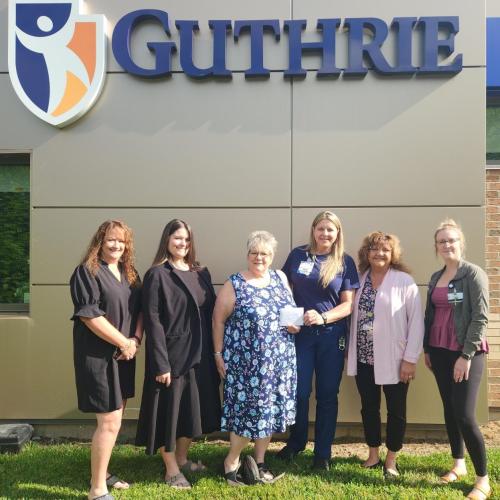 A group of women stand under a Guthrie sign while holding a check.